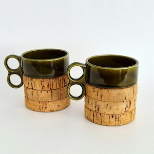 May include: Two ceramic mugs with a glossy dark olive green glaze. Each mug features a distinctive handle with two stacked circular loops. The base of each mug is encased in a thick, textured natural cork sleeve composed of three stacked rings.