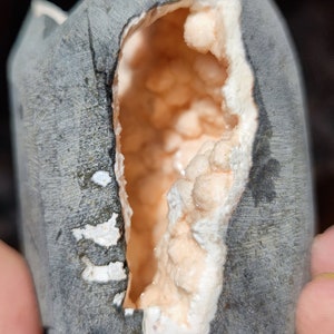 Könnte beinhalten: Ein grauer Stein mit einem gebrochenen Abschnitt, der eine rosa und weiße Kristallgeode im Inneren freilegt.