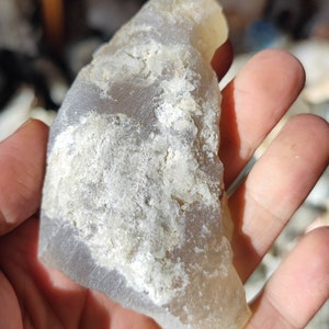 K&ouml;nnte beinhalten: Ein hellgrauer Achat-Stein mit wei&szlig;en B&auml;ndern und einer rauen, strukturierten Oberfl&auml;che.