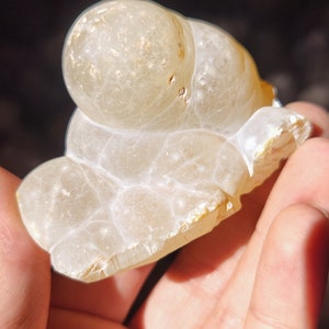 Może przedstawiać: Biała geoda agatowa o gładkiej, zaokrąglonej powierzchni i przezroczystym, mlecznobiałym kolorze. Geoda ma naturalny, organiczny kształt i zawiera kilka małych, brązowych inkluzji.