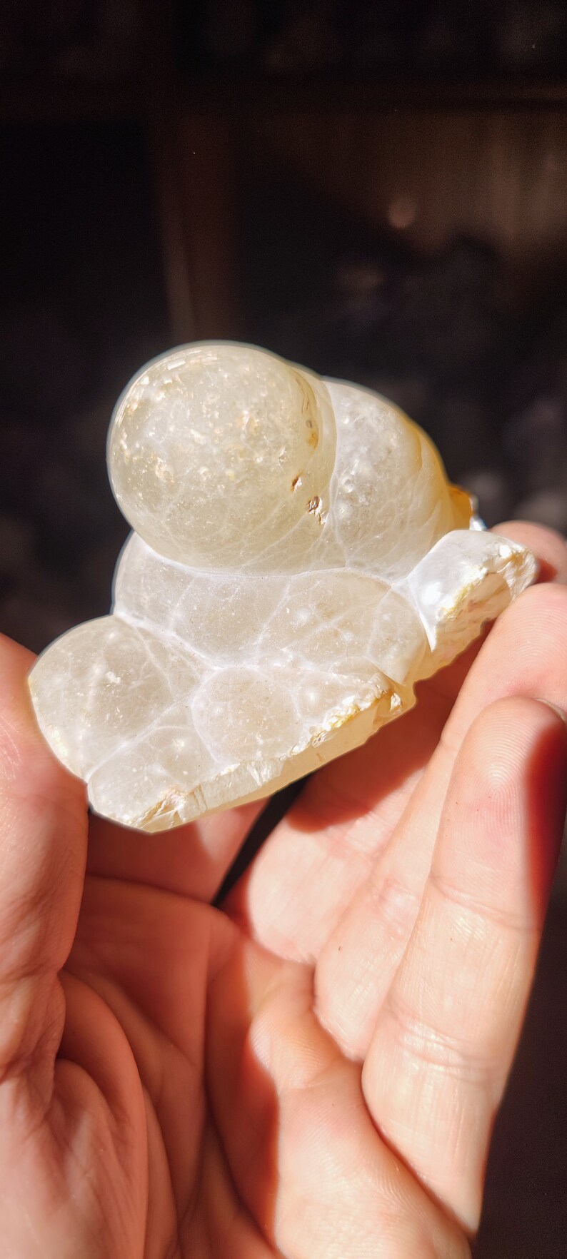 Może przedstawiać: Biała geoda agatowa o gładkiej, zaokrąglonej powierzchni i przezroczystym, mlecznobiałym kolorze. Geoda ma naturalny, organiczny kształt i zawiera kilka małych, brązowych inkluzji.