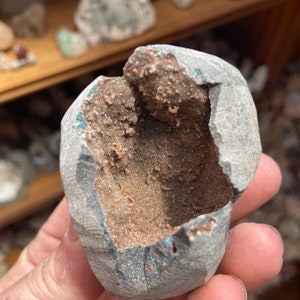 Könnte beinhalten: Ein Geode mit einer gebrochenen, offenen Höhle, die einen braunen, funkelnden, kristallinen Innenraum enthüllt. Der Geode ist grau und hat eine raue, strukturierte Außenseite.