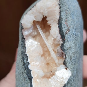 Könnte beinhalten: Eine Geode mit einer rauen, grauen Außenseite und einem Hohlraum, der mit funkelnden, hellfarbigen Kristallen gefüllt ist. Ein langer, dünner Kristall erstreckt sich über die Öffnung. Die Geode wird in einer Hand gehalten.