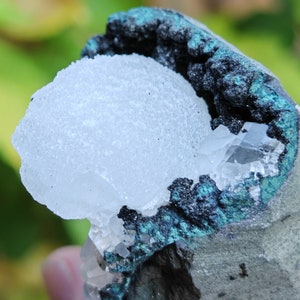 May include: A white crystal cluster with a rough, textured surface is nestled inside a dark grey and teal green rock. The crystal cluster is partially visible, with the rock surrounding it.