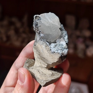 May include: A small, grey and white crystal cluster with a rough, textured surface. The crystal cluster is in the shape of a small, irregular mound.