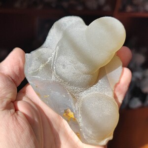 May include: A light gray agate geode with a smooth, rounded surface. The geode has a natural, organic shape and features a few small, yellow inclusions.