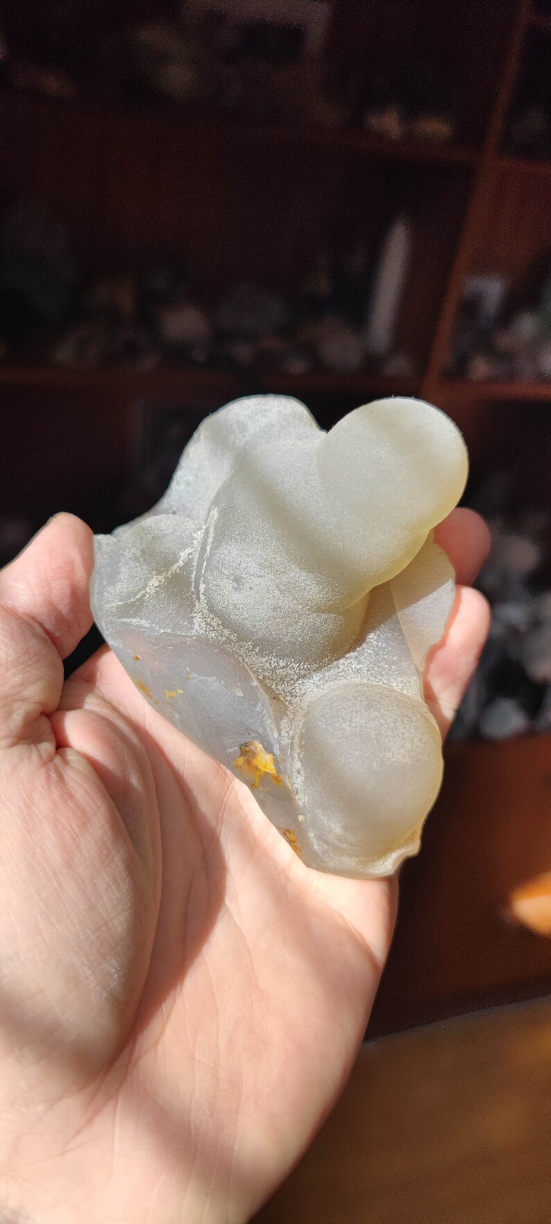 May include: A light gray agate geode with a smooth, rounded surface. The geode has a natural, organic shape and features a few small, yellow inclusions.