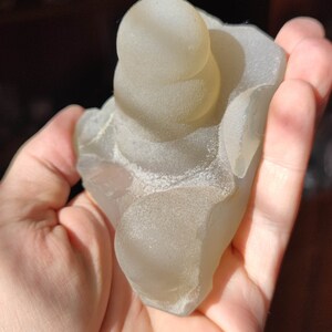 May include: A translucent, light gray, agate geode with a stacked, rounded, smooth, top formation. The geode is in the palm of a hand.