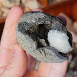 May include: A hand holding a geode. The geode has a dark grey exterior and a cavity filled with white crystalline formations. The outer shell has a rough, textured appearance, and the interior crystals sparkle.