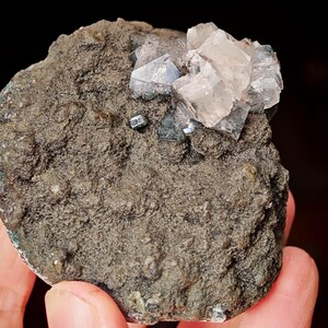 May include: A cluster of clear, translucent crystals with a light pink hue, set in a rough, dark grey rock. The crystals are arranged in a random pattern, with some appearing to be stacked on top of each other. The rock is textured and has a slightly rough surface.