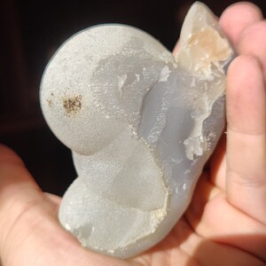 Peut inclure: Une pierre d'agate gris clair avec une surface lisse et arrondie et un bord rugueux et textur&eacute;. La pierre a une forme naturelle et organique et une qualit&eacute; subtile et translucide.