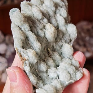 Puede incluir: Esp&eacute;cimen mineral de color azul claro con una superficie texturizada y cristalina. El mineral tiene una apariencia en capas con formaciones redondeadas y agrupadas. El esp&eacute;cimen se sostiene en una mano, mostrando su color natural y estructura &uacute;nica.