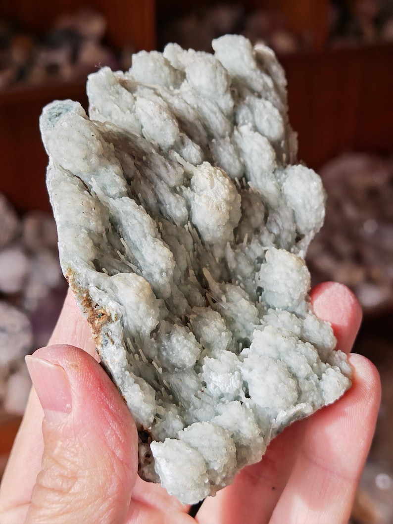 Puede incluir: Esp&eacute;cimen mineral de color azul claro con una superficie texturizada y cristalina. El mineral tiene una apariencia en capas con formaciones redondeadas y agrupadas. El esp&eacute;cimen se sostiene en una mano, mostrando su color natural y estructura &uacute;nica.