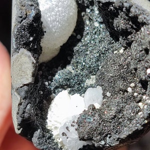May include: Close-up of a geode with a dark grey exterior and a cavity filled with sparkling, small crystals. The interior features white, spherical formations and a dark, granular matrix.