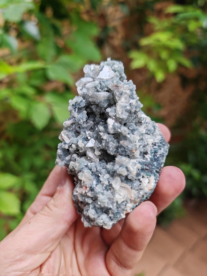 May include: A cluster of light grey and white crystals with a green and grey base. The crystals are clustered together in a rough, natural formation.
