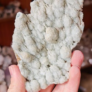 Puede incluir: Un esp&eacute;cimen mineral de color azul verdoso p&aacute;lido con una superficie texturizada y cristalina. El mineral tiene una apariencia agrupada y burbujeante con peque&ntilde;as formaciones redondeadas. El color general es un azul verdoso p&aacute;lido, con algunas &aacute;reas que parecen m&aacute;s blancas. El esp&eacute;cimen se sostiene en una mano.