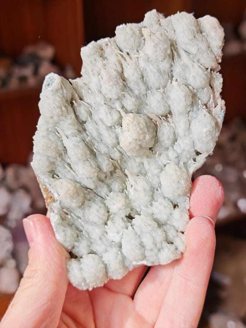 Puede incluir: Un esp&eacute;cimen mineral de color azul verdoso p&aacute;lido con una superficie texturizada y cristalina. El mineral tiene una apariencia agrupada y burbujeante con peque&ntilde;as formaciones redondeadas. El color general es un azul verdoso p&aacute;lido, con algunas &aacute;reas que parecen m&aacute;s blancas. El esp&eacute;cimen se sostiene en una mano.