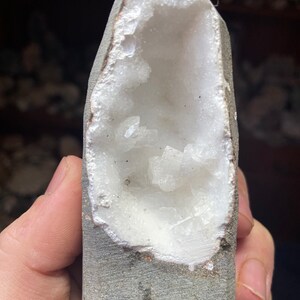 Könnte beinhalten: Ein weißer Geode mit einer großen Öffnung, die einen Cluster weißer Kristalle im Inneren freilegt. Der Geode wird in einer Hand gehalten.