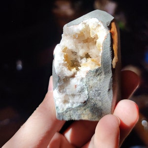 Könnte beinhalten: Eine Geode mit einem rauen, grauen Äußeren und einem hohlen Inneren, das mit funkelnden weißen und gelben Kristallen gefüllt ist. Die Geode wird in einer Hand gehalten und zeigt ihre natürliche Schönheit und einzigartige Struktur.