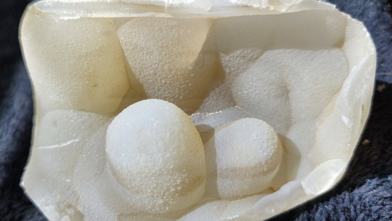 May include: Close-up of a white, round, textured geode with a smooth, white interior. The geode is partially open, revealing the crystal formations inside.