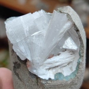 Könnte beinhalten: Ein weißer Kristallcluster, eingebettet in einen grauen Stein. Der Kristall hat eine scharfe, eckige Form und ist durchscheinend.