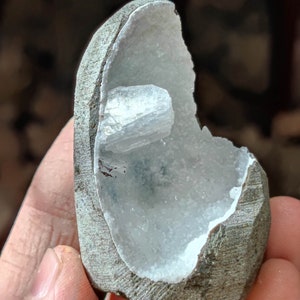 Könnte beinhalten: Eine Geode, eine Gesteinsformation mit einem hohlen Inneren, das mit Kristallen ausgekleidet ist. Die Außenseite ist ein rauer, grauer Stein, während das Innere eine Ansammlung weißer Kristalle zeigt. Eine kleinere Kristallformation ist ebenfalls sichtbar.