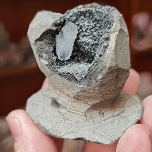 May include: A grey, roughly textured stone sculpture with a hollowed-out centre. The interior features a textured, dark grey surface with a light blue crystal formation. The sculpture has a pedestal base and is held in a person's hand.