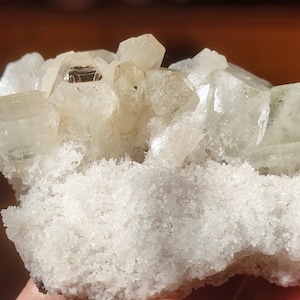 Könnte beinhalten: Ein Cluster aus klaren, weißen und hellgrünen Kristallen auf einer weißen, rauen und strukturierten Basis. Die Kristalle sind in einem Cluster angeordnet, wobei einige der Kristalle nach oben zeigen.