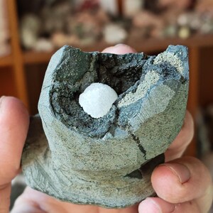 May include: A hand-held, rough-textured, grey-green rock formation with a central cavity. Inside the cavity is a small, spherical, white crystal cluster. The rock has a layered appearance, with areas of darker grey and lighter green.