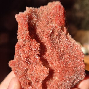 Könnte beinhalten: Nahaufnahme eines roten, kristallinen Gesteins mit einer rauen, strukturierten Oberfläche. Der Stein wird in einer Hand gehalten, und das Licht fängt die Kristalle ein, wodurch sie funkeln.