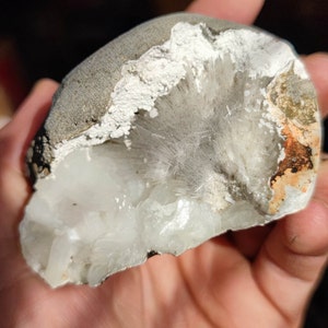 Puede incluir: Primer plano de una roca gris con un cristal blanco en su interior. El cristal tiene una textura fibrosa y está parcialmente expuesto.