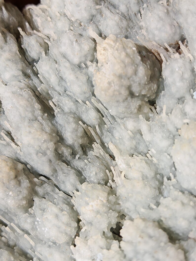 Puede incluir: Primer plano de una formaci&oacute;n mineral azul claro con una superficie texturizada. El mineral tiene una estructura cristalina con peque&ntilde;os grupos redondeados y finas protuberancias en forma de aguja, creando una apariencia org&aacute;nica &uacute;nica.