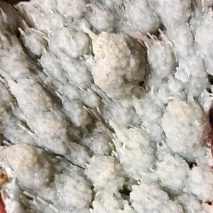 Puede incluir: Primer plano de una formaci&oacute;n mineral gris claro con una apariencia texturizada y cristalina. La superficie est&aacute; cubierta de peque&ntilde;os grupos redondeados y estructuras en forma de aguja, creando un patr&oacute;n natural &uacute;nico. El color general es un gris p&aacute;lido y suave.