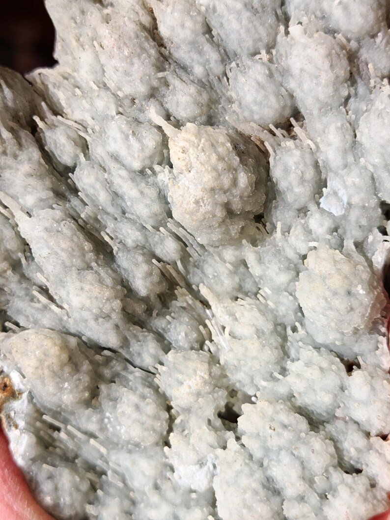 Puede incluir: Primer plano de una formaci&oacute;n mineral gris claro con una apariencia texturizada y cristalina. La superficie est&aacute; cubierta de peque&ntilde;os grupos redondeados y estructuras en forma de aguja, creando un patr&oacute;n natural &uacute;nico. El color general es un gris p&aacute;lido y suave.