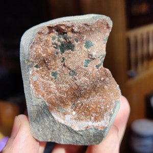 Könnte beinhalten: Nahaufnahme eines Steins mit einer abgebrochenen Kante, die einen kristallgefüllten Innenraum freilegt. Die Kristalle sind eine Mischung aus Weiß und Braun, mit einigen grünen und schwarzen Einschlüssen. Der Stein wird in einer Hand gehalten.