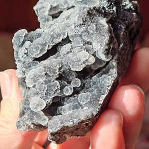 May include: A cluster of gray and black chalcedony with a bubbly texture. The chalcedony is translucent and has a light blue hue.