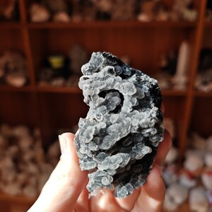 May include: A close-up of a gray and black crystal with a unique, bubbly texture. The crystal is being held in a hand.