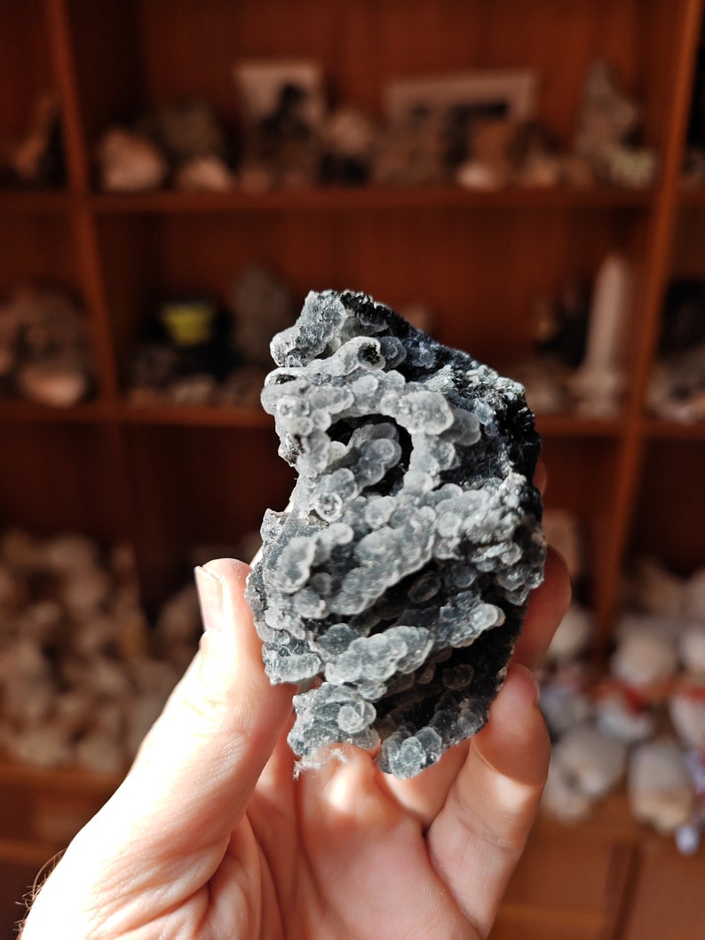 May include: A close-up of a gray and black crystal with a unique, bubbly texture. The crystal is being held in a hand.