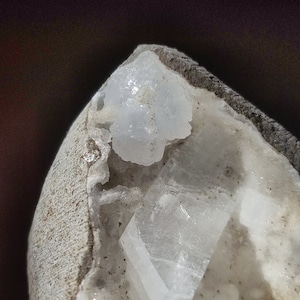 May include: A close-up of a white crystal cluster in a grey rock. The crystal is translucent and has a sharp, geometric shape.