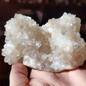 Könnte beinhalten: Ein Cluster aus klaren Quarz Kristallen. Die Quarzformation hat eine helle, cremeweiße Farbe und wird in einer Hand gehalten. Die Kristalle haben eine natürliche, raue Textur und sind zusammengefasst. Etwa 10 cm lang.