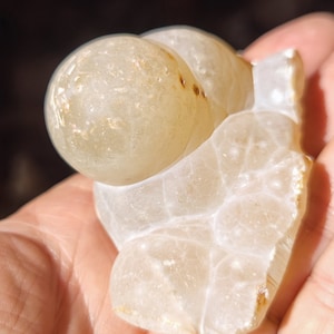 Może przedstawiać: Biała geoda agatowa z przezroczystym, mlecznobiałym wnętrzem i brązowymi oznaczeniami. Geoda ma kształt serca i gładką, polerowaną powierzchnię.