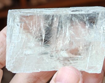 May include: A clear, translucent, rectangular crystal with a striated surface. The crystal is approximately 2 inches long and 1 inch wide.