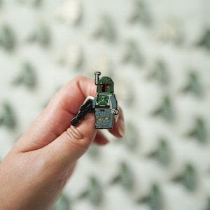 Pode incluir: Um broche de esmalte de Boba Fett, um personagem de Star Wars, segurando um blaster. O broche tem a forma de uma minifigura Lego e é verde e preto.