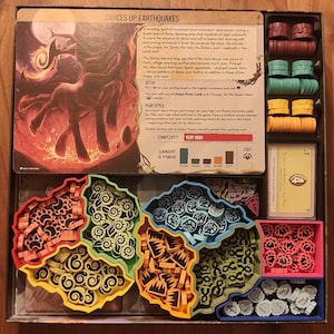 May include: A board game box with various components. The box contains cards, tokens, and organizers in multiple colors, including red, yellow, green, and blue. The game's title, "Dances Up Earthquakes," is visible on the box's interior.