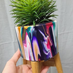 May include: A colourful, abstractly painted ceramic planter with a wooden stand. The planter has a swirling pattern of purple, pink, white, gold, and green paint. A small green succulent plant is in the planter.