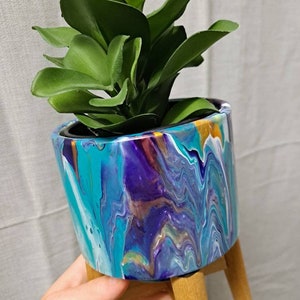 May include: A small plant in a colourful, abstract, blue, purple and white painted planter with a wooden stand.