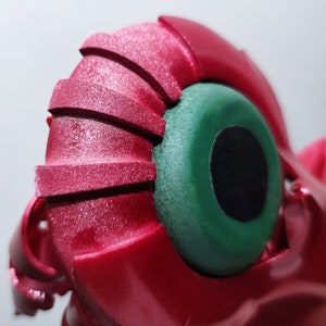 以下が含まれることがあります： テクスチャ加工された表面を持つ赤と緑のロボットの頭部のクローズアップ。ロボットは、黒い中心を持つ大きく丸い緑色の目をしています。