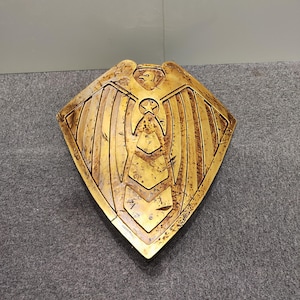 Op de afbeelding: Een gouden schild met een gestileerd adelaarontwerp en een ster in het midden. Het schild heeft een verweerde, oude uitstraling.
