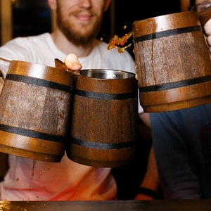 May include: Three wooden mugs with black bands around the middle are being clinked together. The mugs are filled with a dark liquid.
