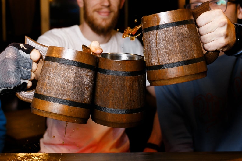 May include: Three wooden mugs with black bands around the middle are being clinked together. The mugs are filled with a dark liquid.
